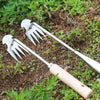 A pair of stainless steel weeding tools with wooden handles are shown lying on dirt and green plants.