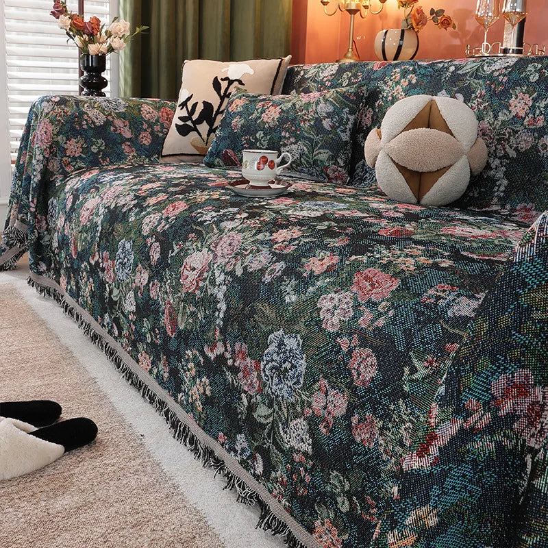 A dark floral jacquard sofa blanket featuring pink and green flowers and decorative tassels on a bed.
