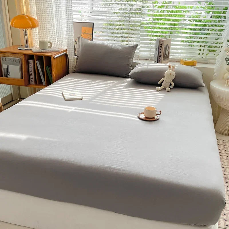 A light gray fitted sheet is laid out on a bed with pillows and a stuffed animal near a window.