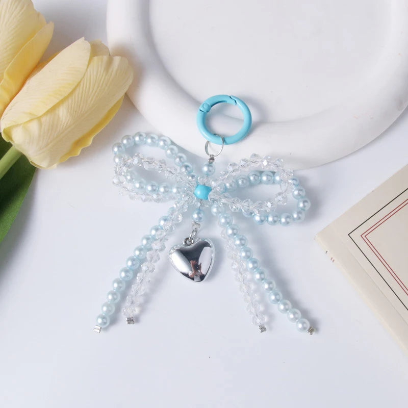 A light blue pearl bow keychain with a silver heart charm resting on a white surface next to a yellow tulip.