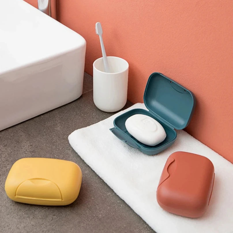 A compact, leak-proof travel soap box in terracotta and yellow colors rests on a white towel next to a toilet.