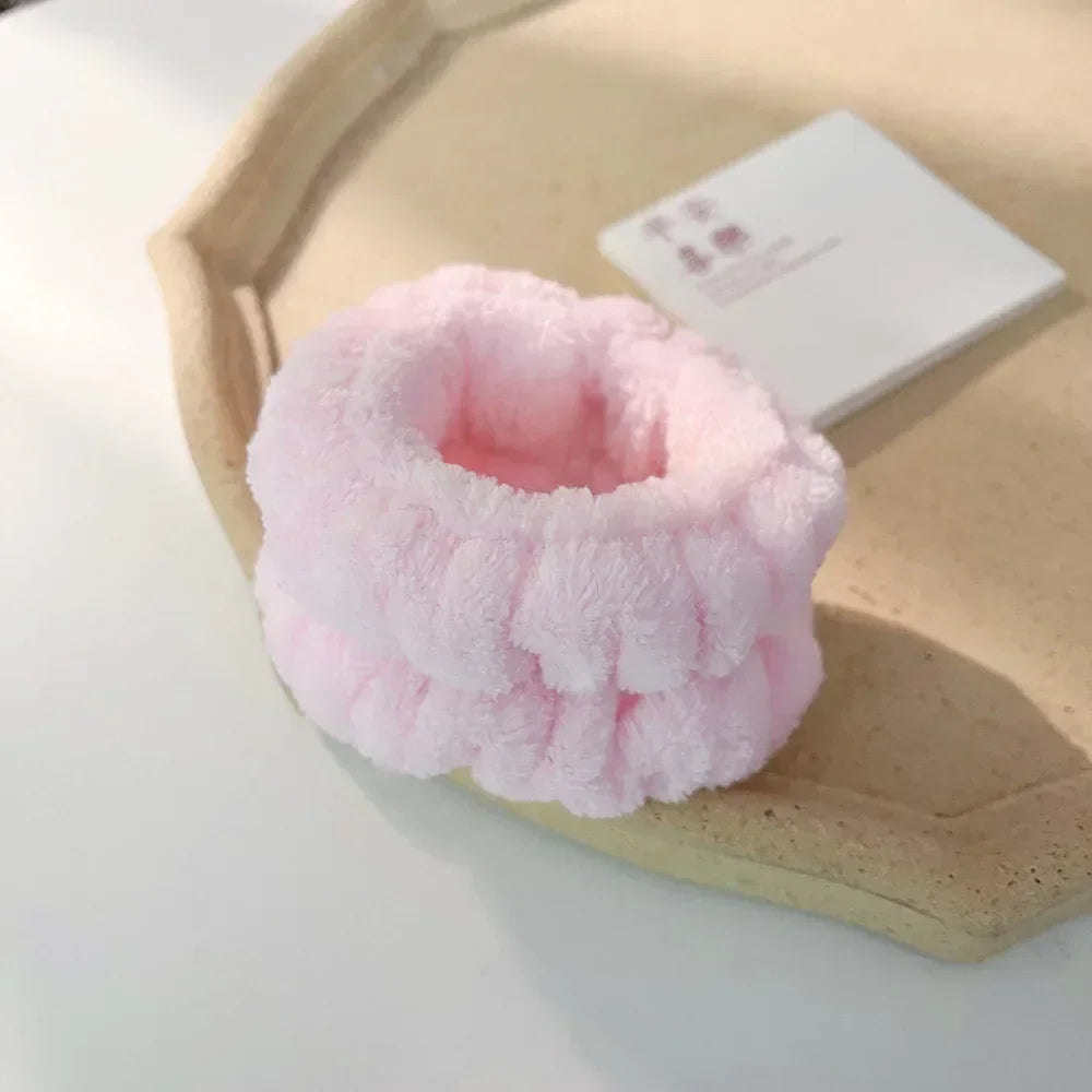 A pink microfiber wrist sweatband and scrunchies for face washing resting on a light wooden tray.