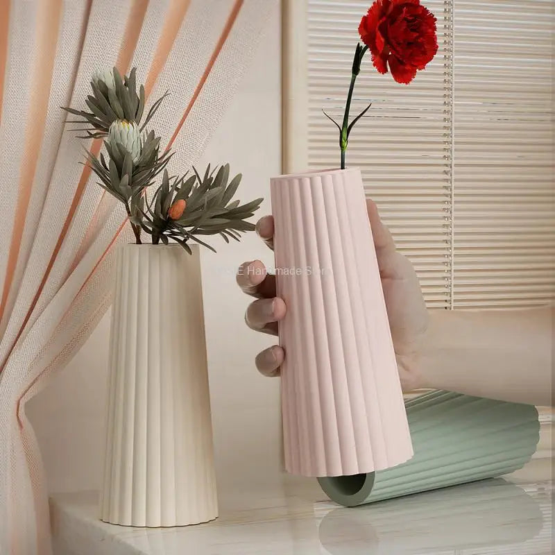 A hand holds a pink silicone mold for candle and vase projects next to a tall white ceramic vase and a red flower.