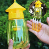 A hand holds a yellow and green glass fruit fly trap kit with pheromone lures against a blurred green background.