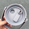 A hand holds a gray insulated stainless steel tumbler with a handle and straw.