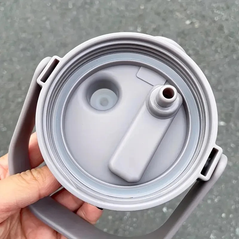 A hand holds a gray insulated stainless steel tumbler with a handle and straw.