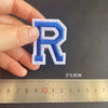 A hand holds a blue embroidered letter R patch against a ruler showing its size.