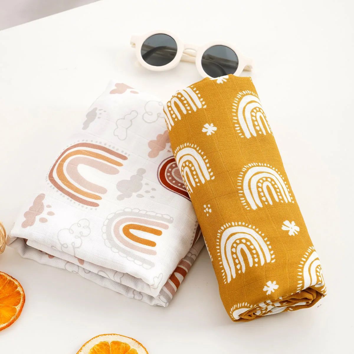 A yellow muslin baby swaddle blanket rolled up next to a pair of sunglasses and an orange slice on a white surface.