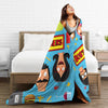 A woman is relaxing on a bed with a colorful cartoon-themed throw blanket featuring characters and text.