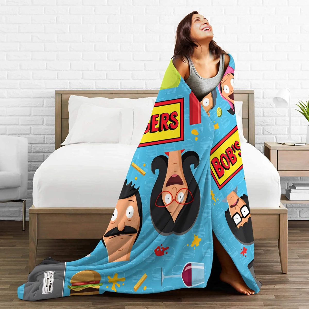 A woman is relaxing on a bed with a colorful cartoon-themed throw blanket featuring characters and text.