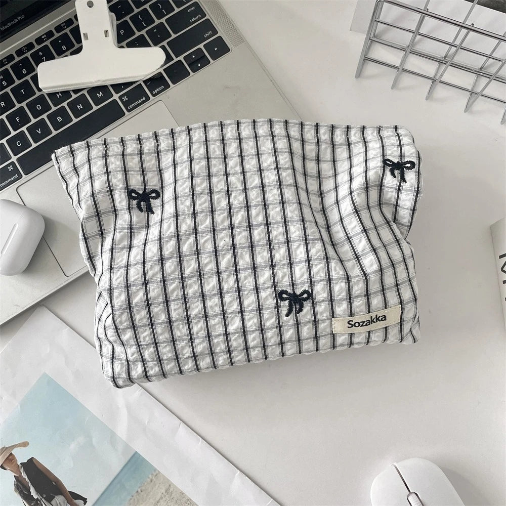 A gray and white striped fabric makeup bag with black floral embroidery rests on a white desk next to a laptop.