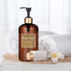 A brown glass body wash bottle with a gold pump dispenser sits on a white surface next to a white towel and a white soap bar.