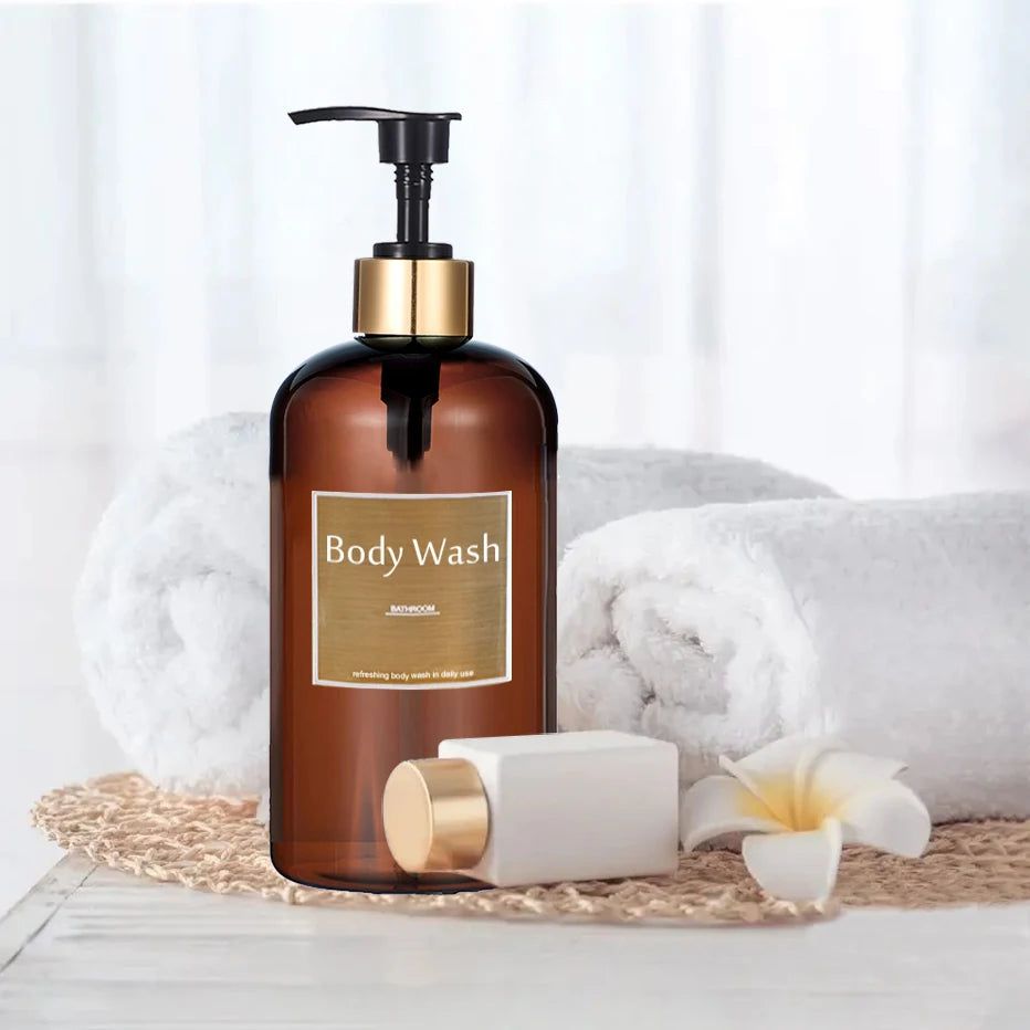 A brown glass body wash bottle with a gold pump dispenser sits on a white surface next to a white towel and a white soap bar.