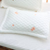 A white quilted baby pillow with small peach-colored embroidered shapes resting on a white textured surface.