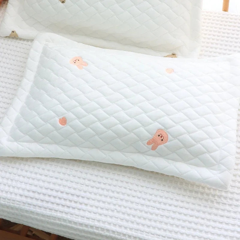 A white quilted baby pillow with small peach-colored embroidered shapes resting on a white textured surface.
