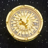 A round, gold-colored metal roulette dice with numbered markings and a central pivot.