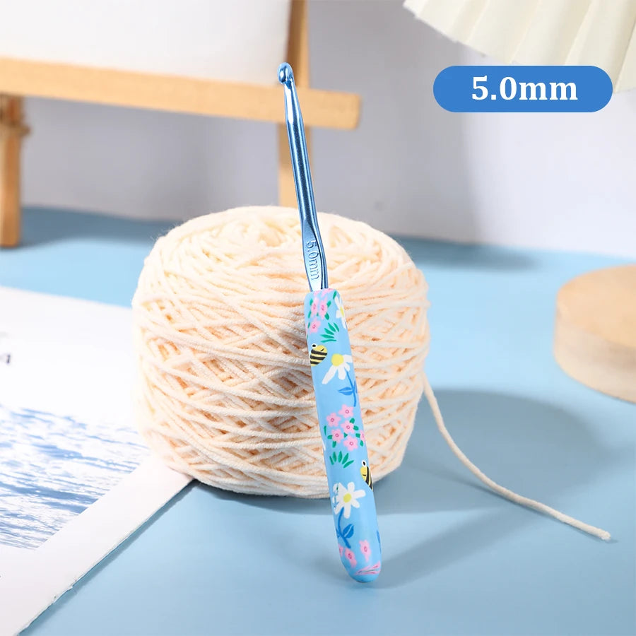 A blue ergonomic crochet hook with a bee design is shown next to a ball of light-colored yarn.