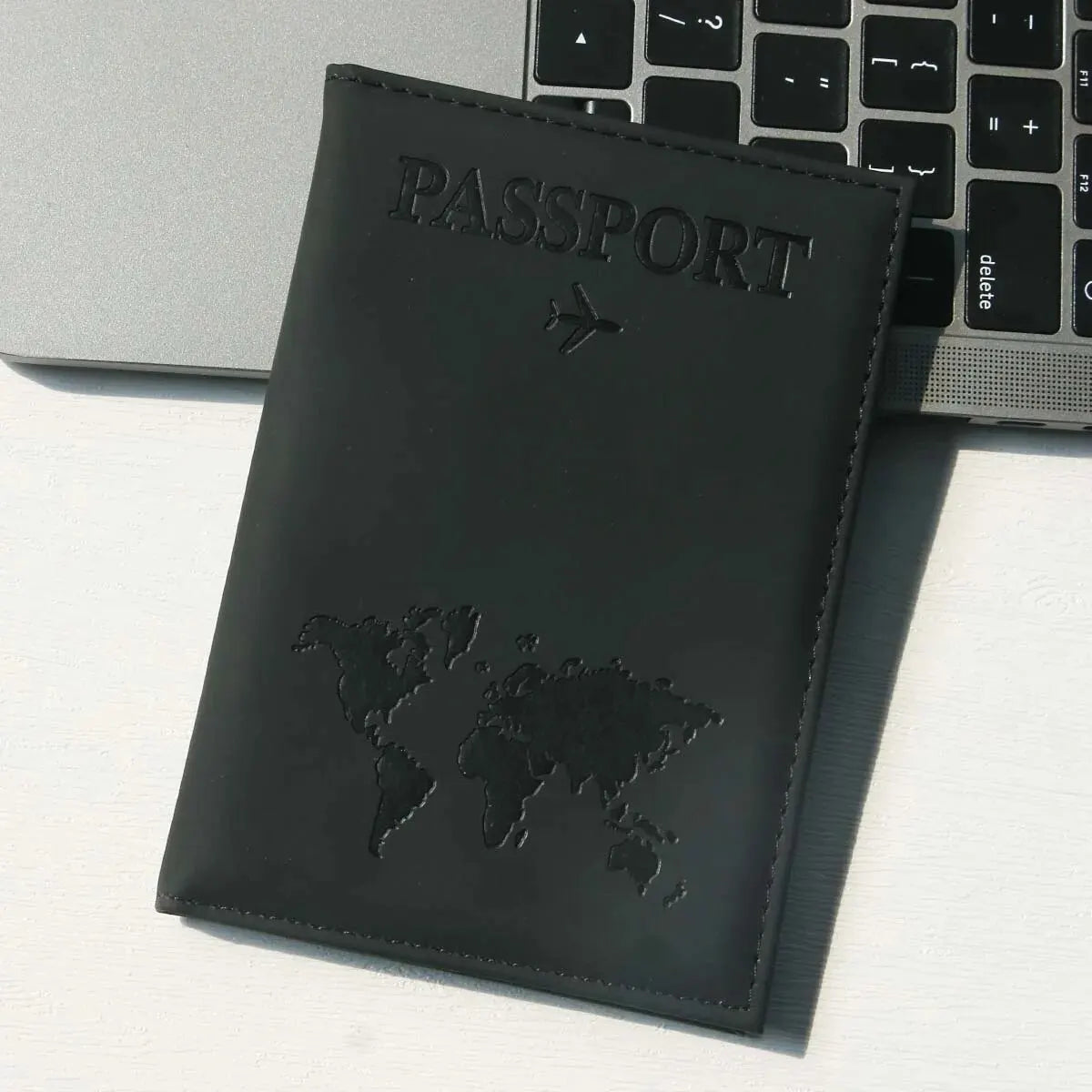 Schwarzes Leder-Reisepass-Etui mit Weltkarten-Design auf weißer Oberfläche neben einem Laptop.