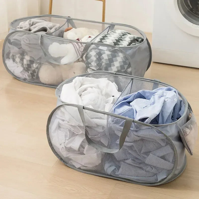A gray folding mesh laundry basket with handles is shown holding folded clothes on a wooden floor.