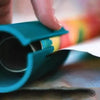 A hand is operating a teal compact safe paper cutter for wrapping and crafts on a surface.