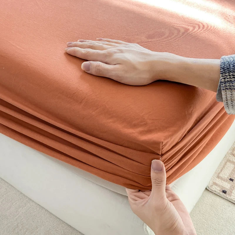 A hand is folding a rust-colored cotton fitted sheet with an elastic band on a white surface.