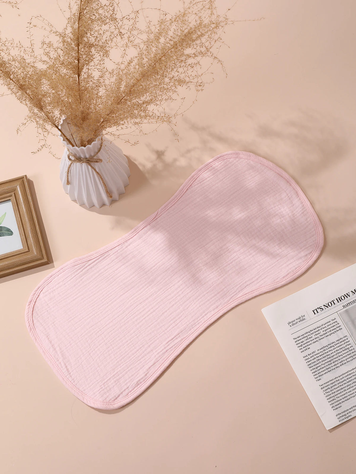 A pink baby burp cloth is displayed on a light background next to dried floral arrangements and a picture frame.