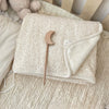 A beige floral muslin baby blanket with a light tan wooden moon-shaped pacifier resting on it.