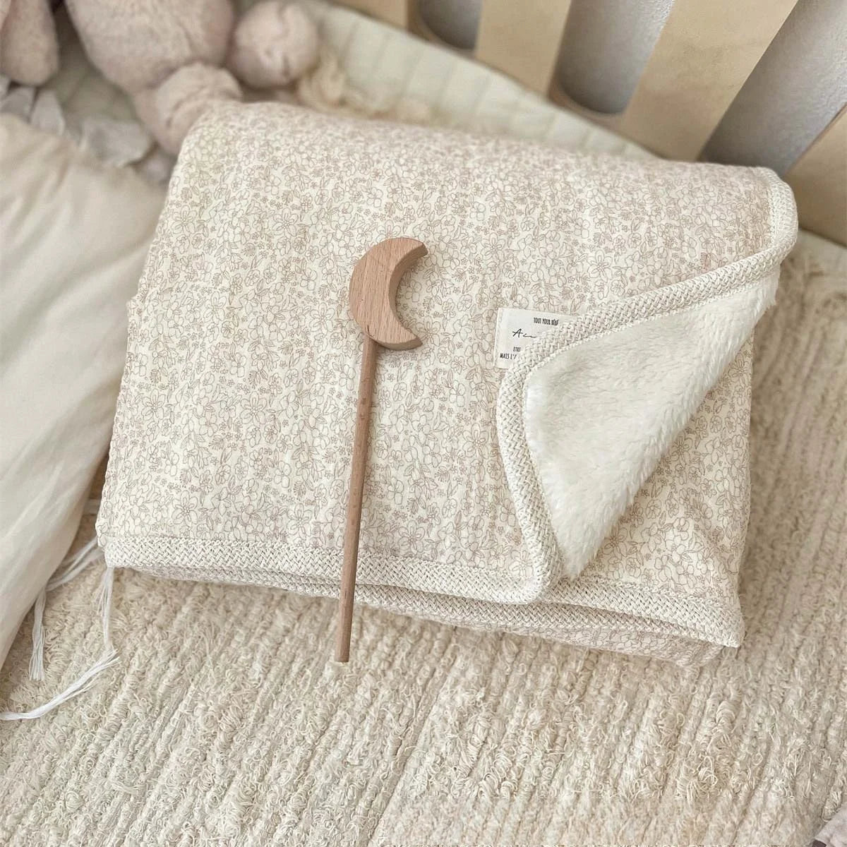 A beige floral muslin baby blanket with a light tan wooden moon-shaped pacifier resting on it.