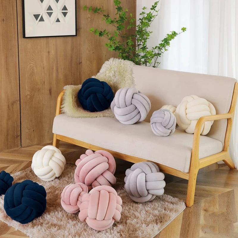 A collection of soft, round knitted velvet throw pillows in shades of gray, pink, and cream displayed on a light-colored sofa