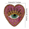 A heart-shaped embroidered patch featuring a large eye design with gold and iridescent beads on a purple fabric background.