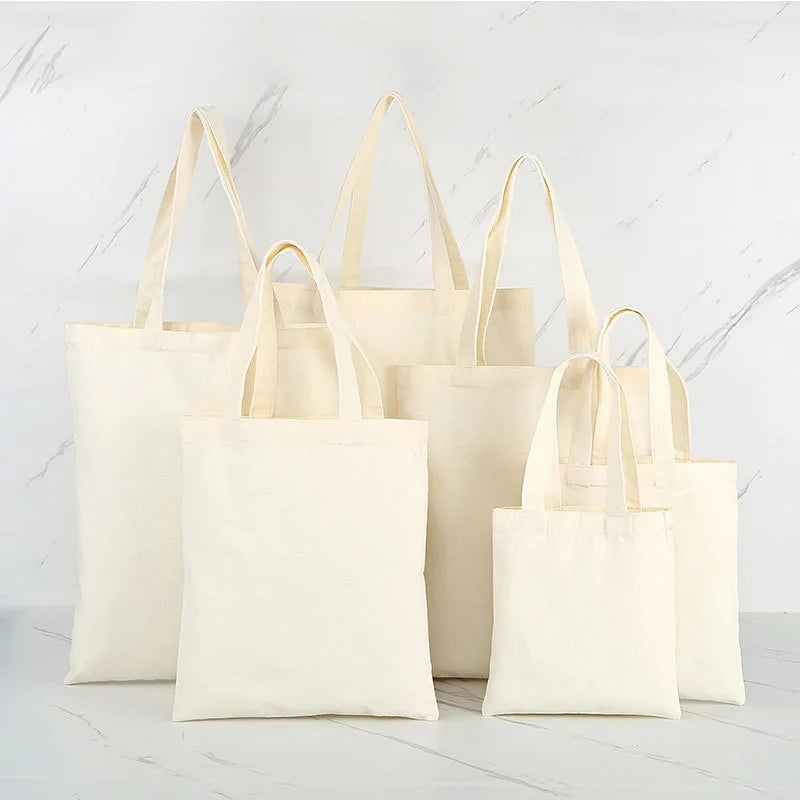 A collection of natural canvas shopping tote bags in off-white and cream colors displayed on a white marble surface.