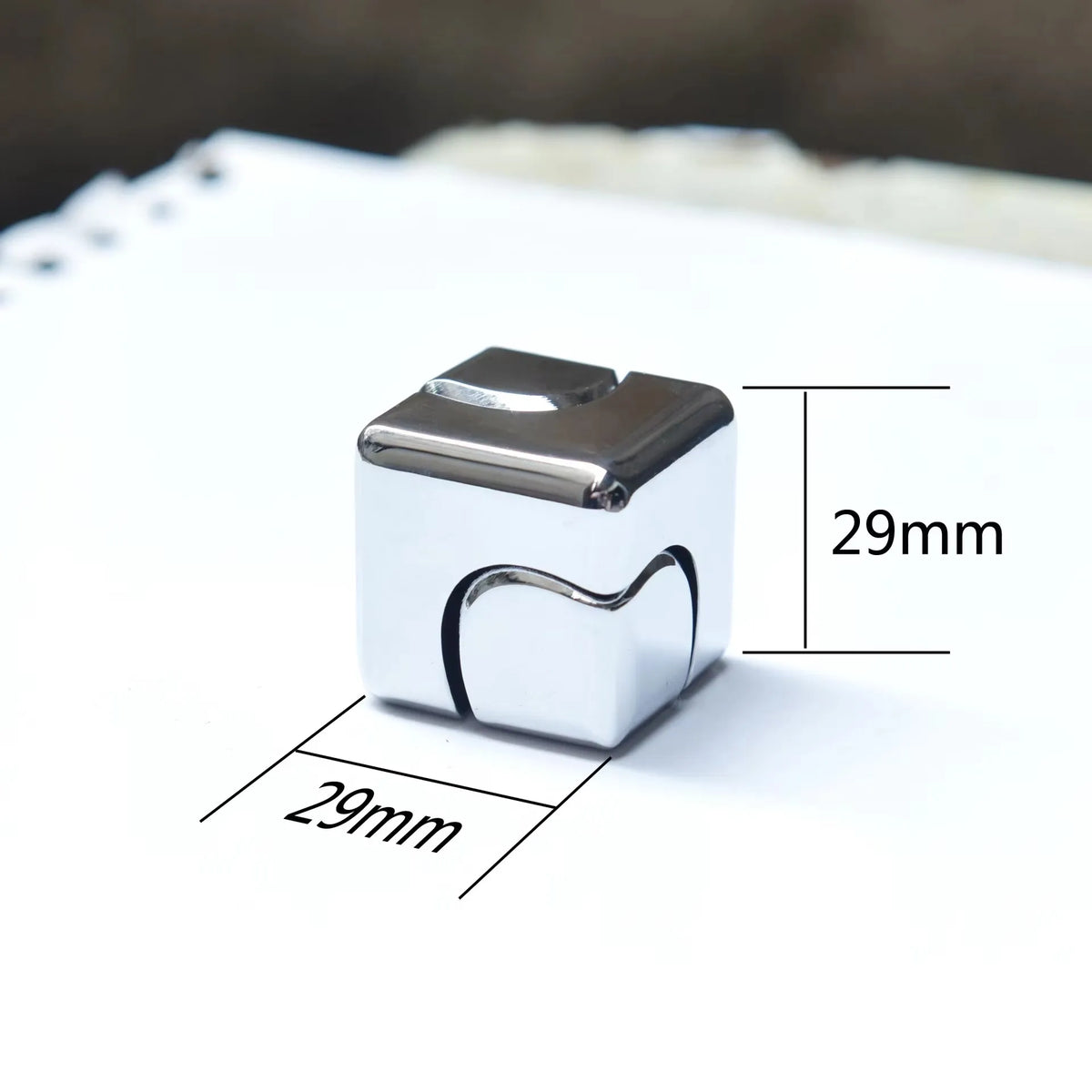 A silver metal fidget spinner cube resting on a white surface with dimension markings.