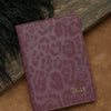 A deep purple leopard print passport holder with gold name embossing resting on a brown surface.