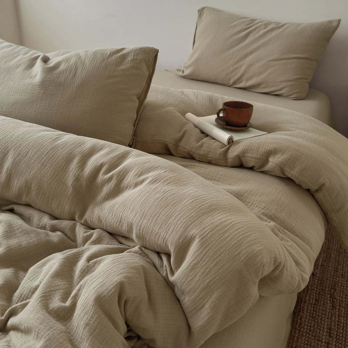 Beige, faltige Bettwäsche-Decke und Kissen sowie eine kleine braune Tasse auf einem Bett.