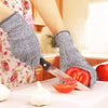 A hand wearing gray cut-resistant work gloves is preparing to cut a tomato and a garlic bulb on a white cutting board.