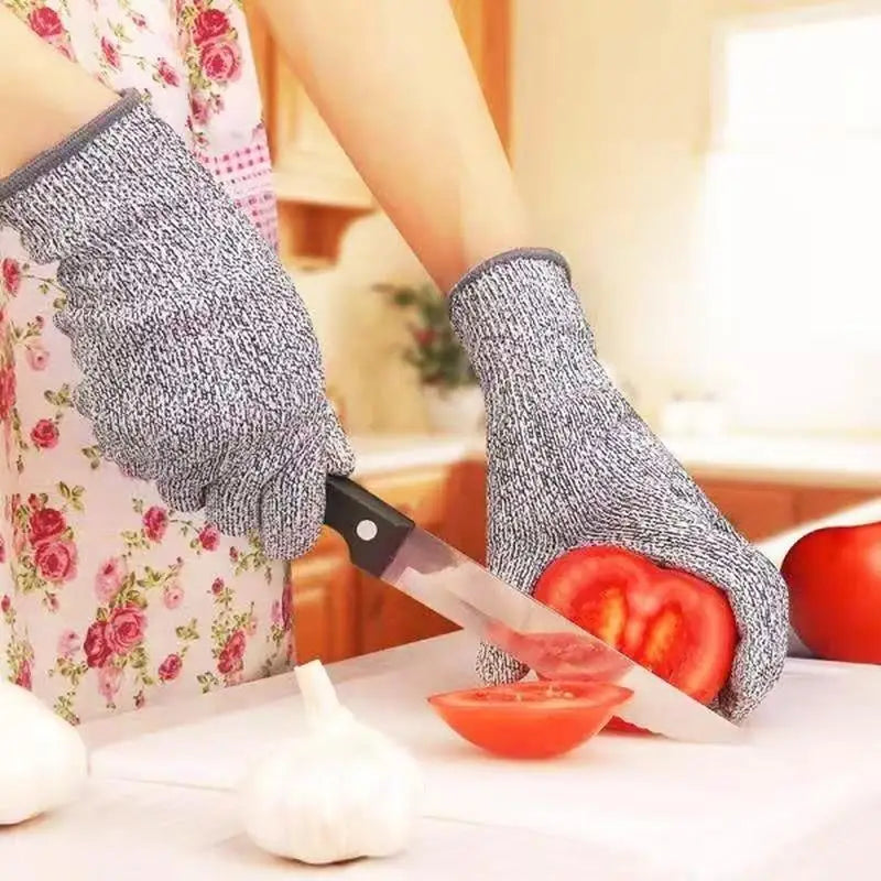 A hand wearing gray cut-resistant work gloves is preparing to cut a tomato and a garlic bulb on a white cutting board.