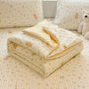 A beige lace-trimmed breathable summer duvet folded on a light patterned bedspread with a teddy bear nearby.