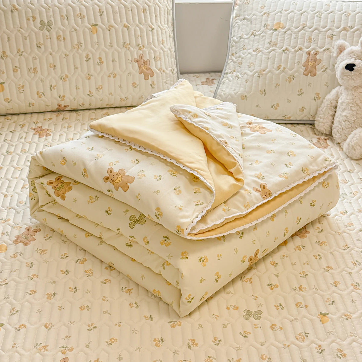 A beige lace-trimmed breathable summer duvet folded on a light patterned bedspread with a teddy bear nearby.