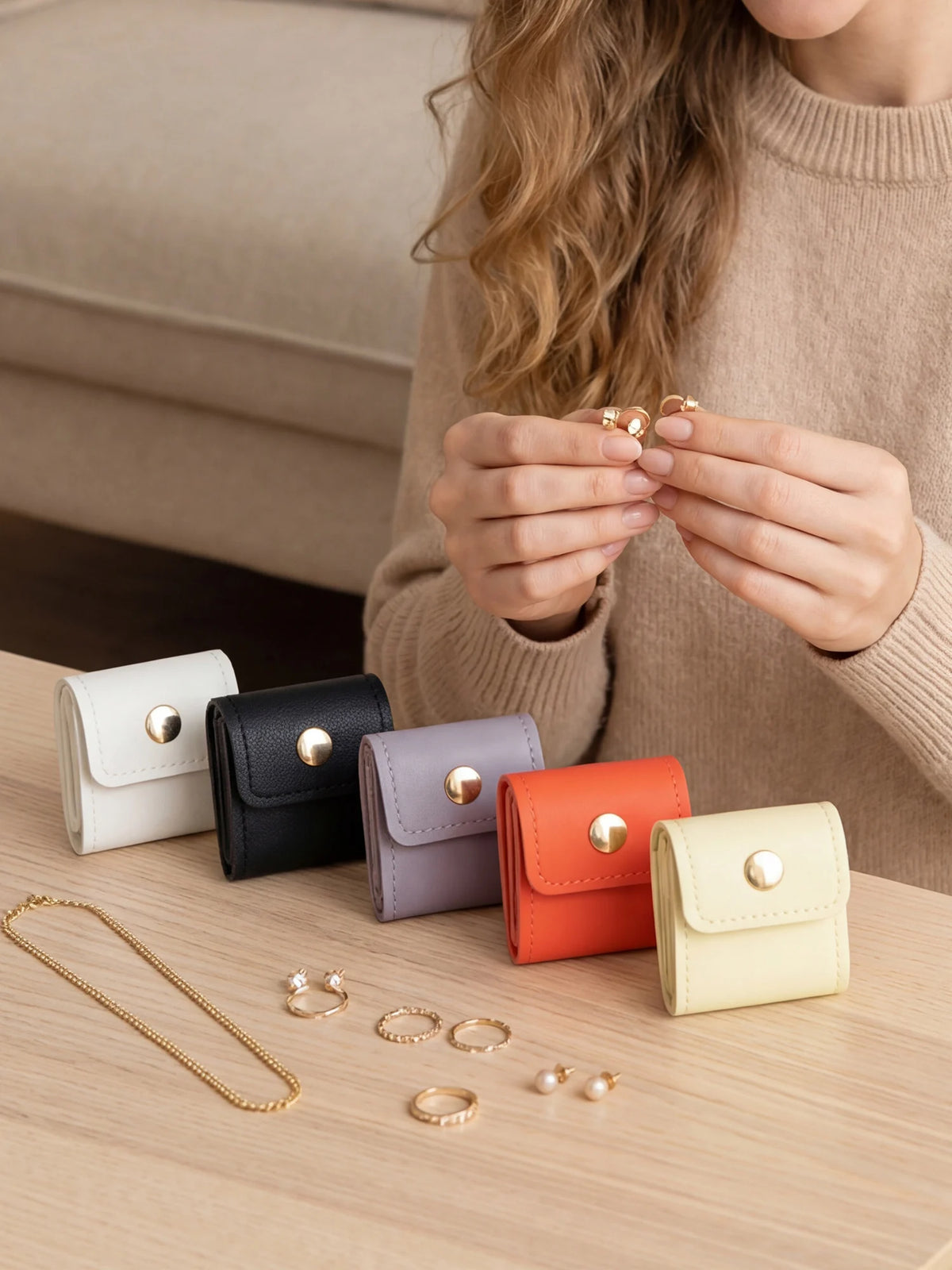 A person is holding a beige sweater and displaying various gold and silver jewelry organizers and pouches on a wooden surface
