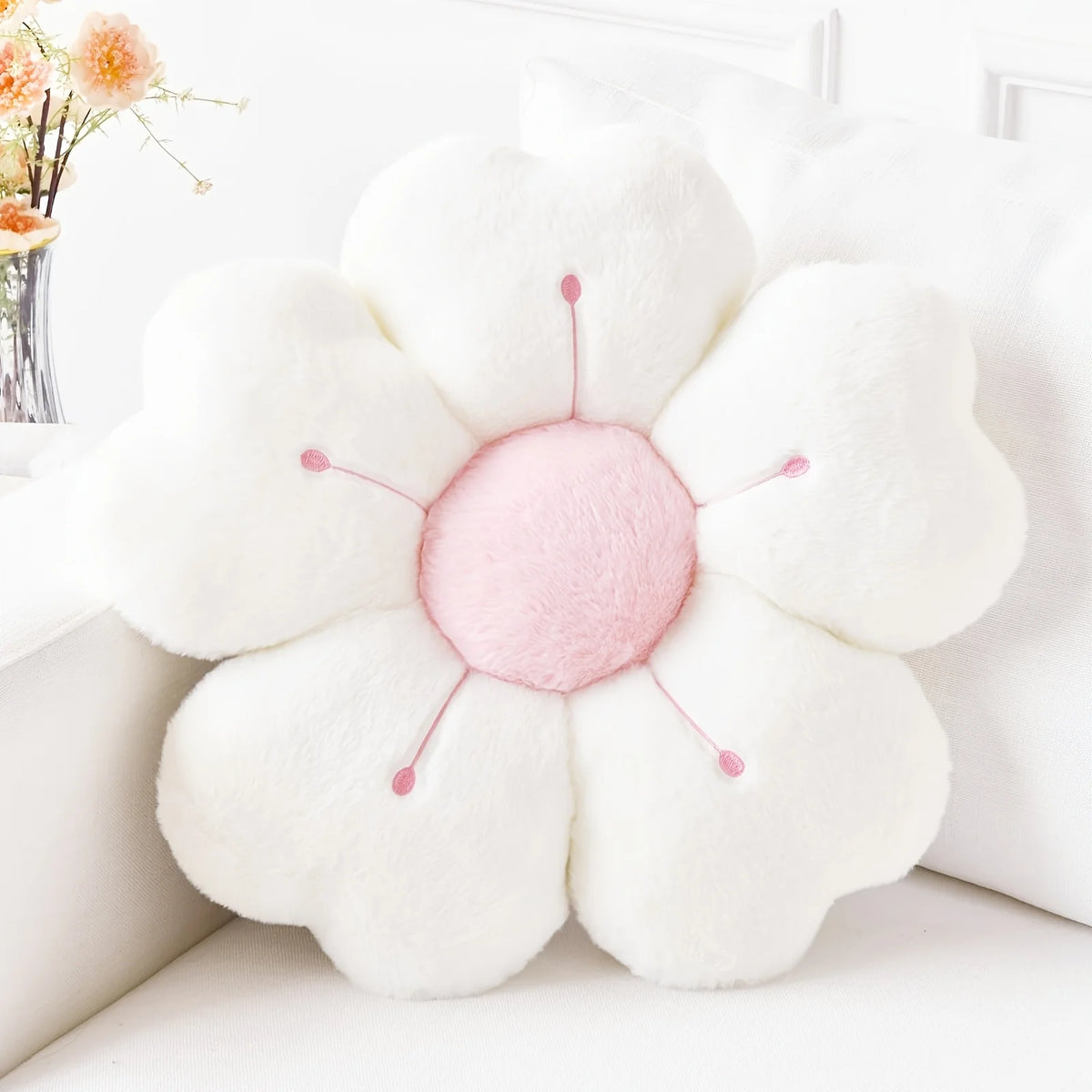 A white plush daisy flower throw pillow with pink center and green stems resting on a white surface.
