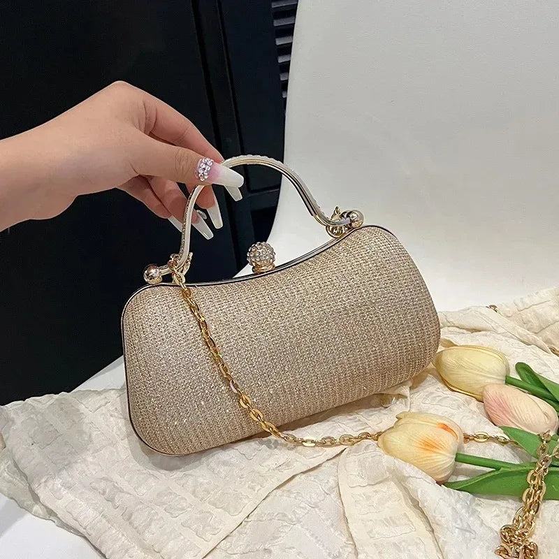 A hand holds a shimmering beige envelope clutch with a gold chain strap on a white textured surface.