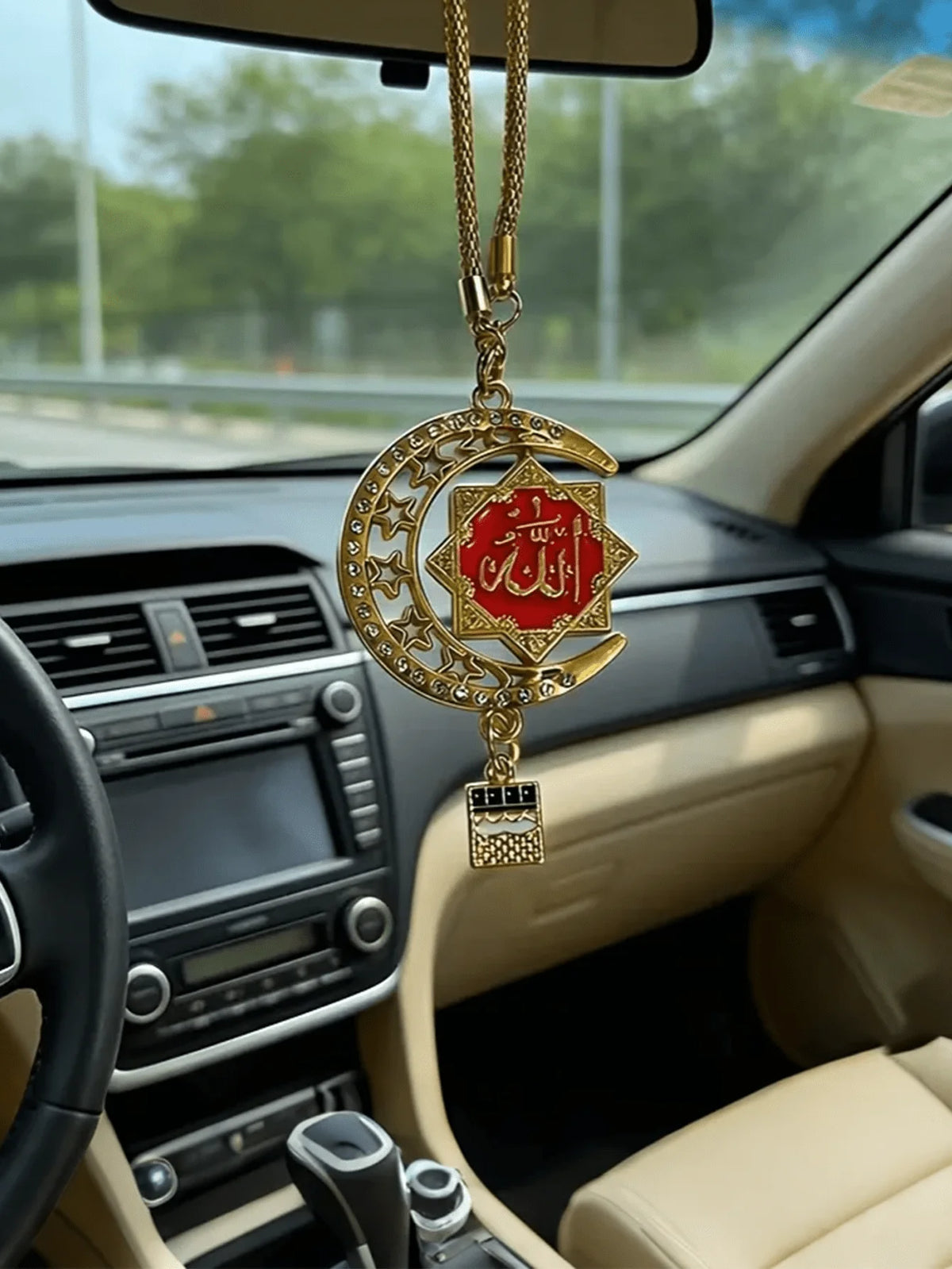 A gold-tone Islamic calligraphy pendant hangs from a chain, displayed inside a car interior.