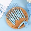 A wooden board holds a set of ergonomic crochet hooks with blue and white bee designs on a light blue background.