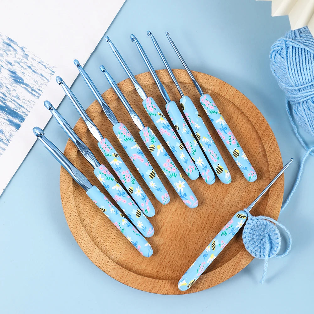 A wooden board holds a set of ergonomic crochet hooks with blue and white bee designs on a light blue background.