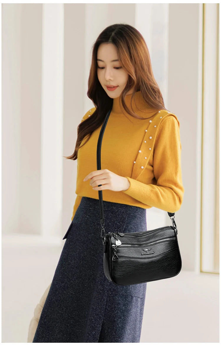 A woman wearing a mustard yellow turtleneck sweater and dark skirt holds a black compact square crossbody bag.