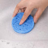 A hand is squeezing a bright blue compressed natural sponge into soapy water.