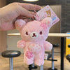 A pink cherry blossom plush bear keychain is held by a hand against a blurred background.