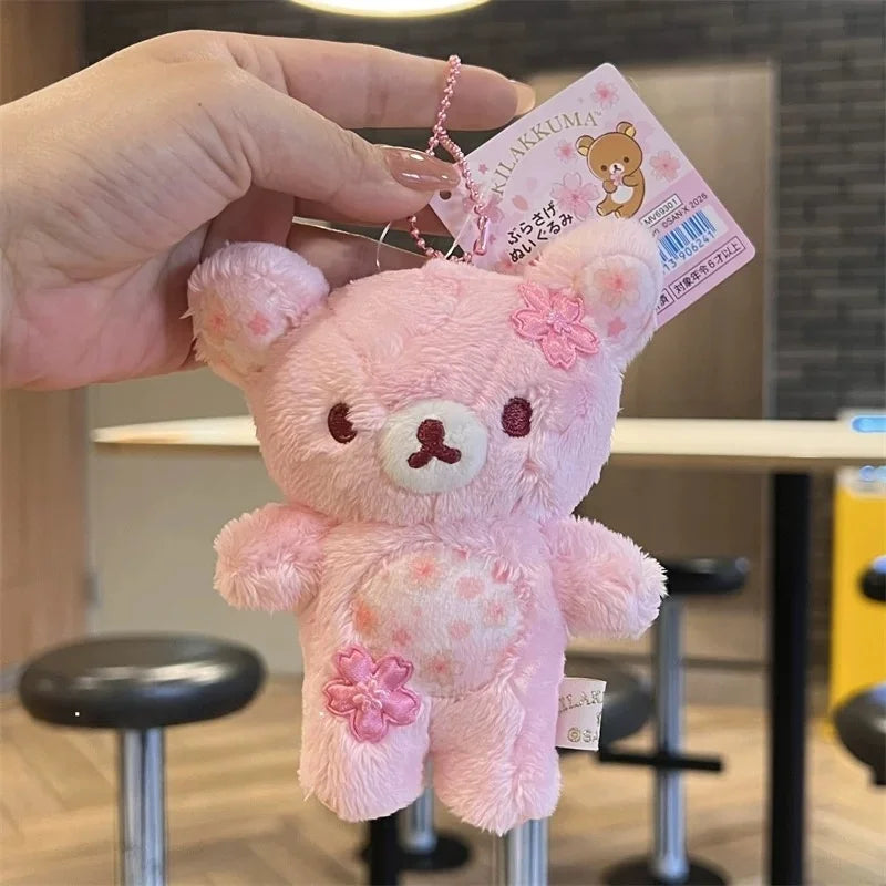 A pink cherry blossom plush bear keychain is held by a hand against a blurred background.