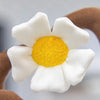 A white artificial flower plush doll with a yellow center is shown against a blurred background.
