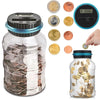 A clear glass piggy bank filled with coins, next to various gold and silver coins and a digital coin counting device.